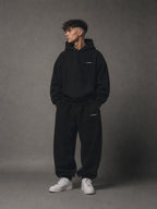 Man wearing a black hoodie and pants with a brand logo on a gray background
