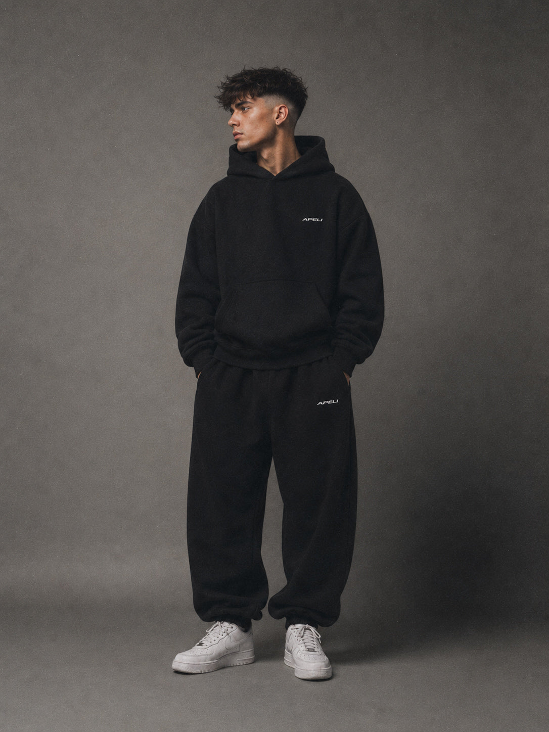 Man wearing a black hoodie and pants with a brand logo on a gray background